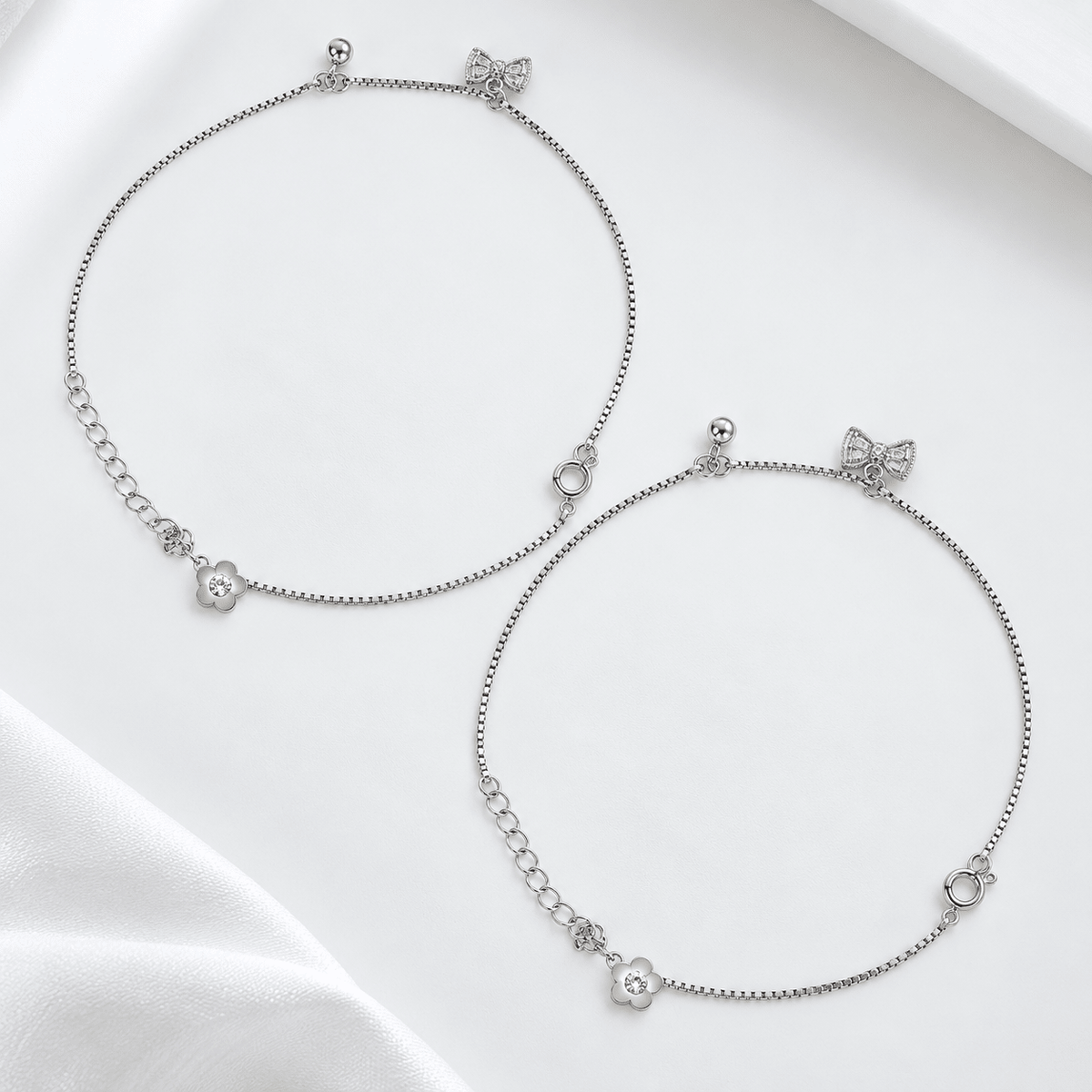 Two delicate silver anklets with bow and flower charms, displayed on a clean white fabric background.