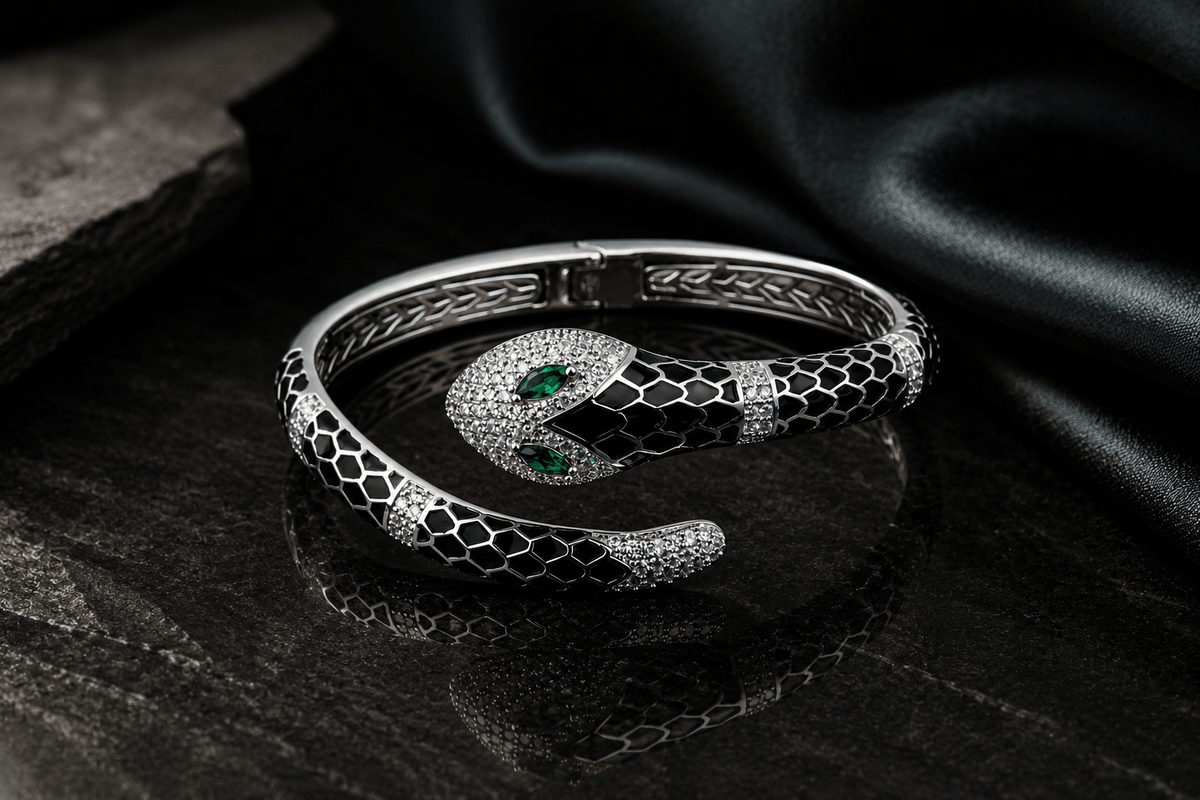 A 925 silver snake-shaped cuff bracelet featuring black enamel scales, pavé-set clear stones, and two green emerald-cut gemstones in the eyes by Miraura Jewels.