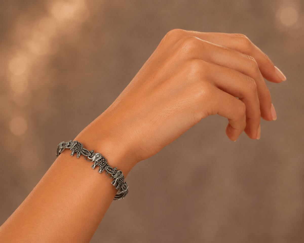 A close-up of a person's wrist wearing a silver-toned chain bracelet with small dangling Elephant charms, set against a softly blurred warm brown background.