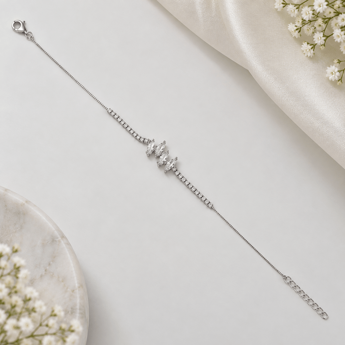 A delicate silver chain bracelet with a central cluster of clear cubic zirconia stones, displayed on a light gray surface with white satin and baby's breath flowers.