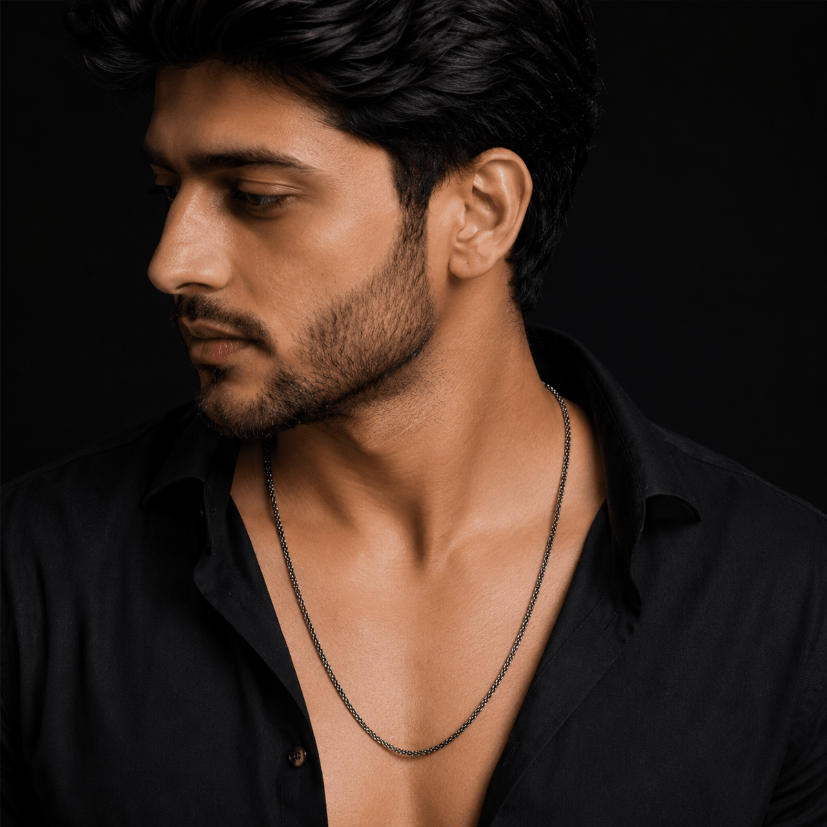 A close-up portrait of a man wearing a dark metallic chain necklace over a black shirt against a black background.