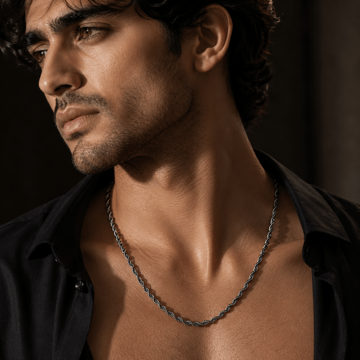 A close-up of a man wearing a oxidised 925 silver twisted rope chain, by miraura jewels styled with an open black shirt against a dark background.