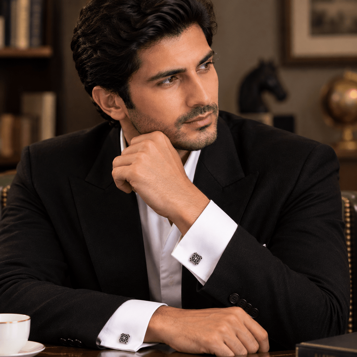 A man in a black suit and white dress shirt resting his chin on his hand, showcasing decorative silver cufflinks on his cuffs, set in a warm, elegant indoor environment.