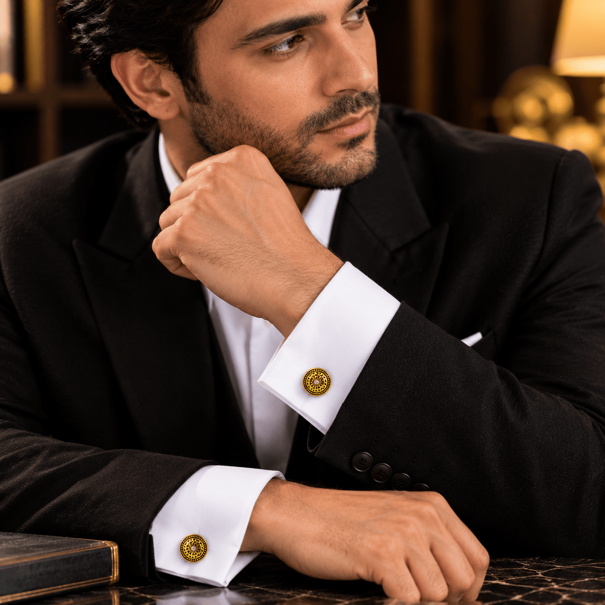 A man in a black suit and white dress shirt wearing round gold cufflinks with a circular emblem, resting his chin on his hand in a warm, elegant indoor setting.