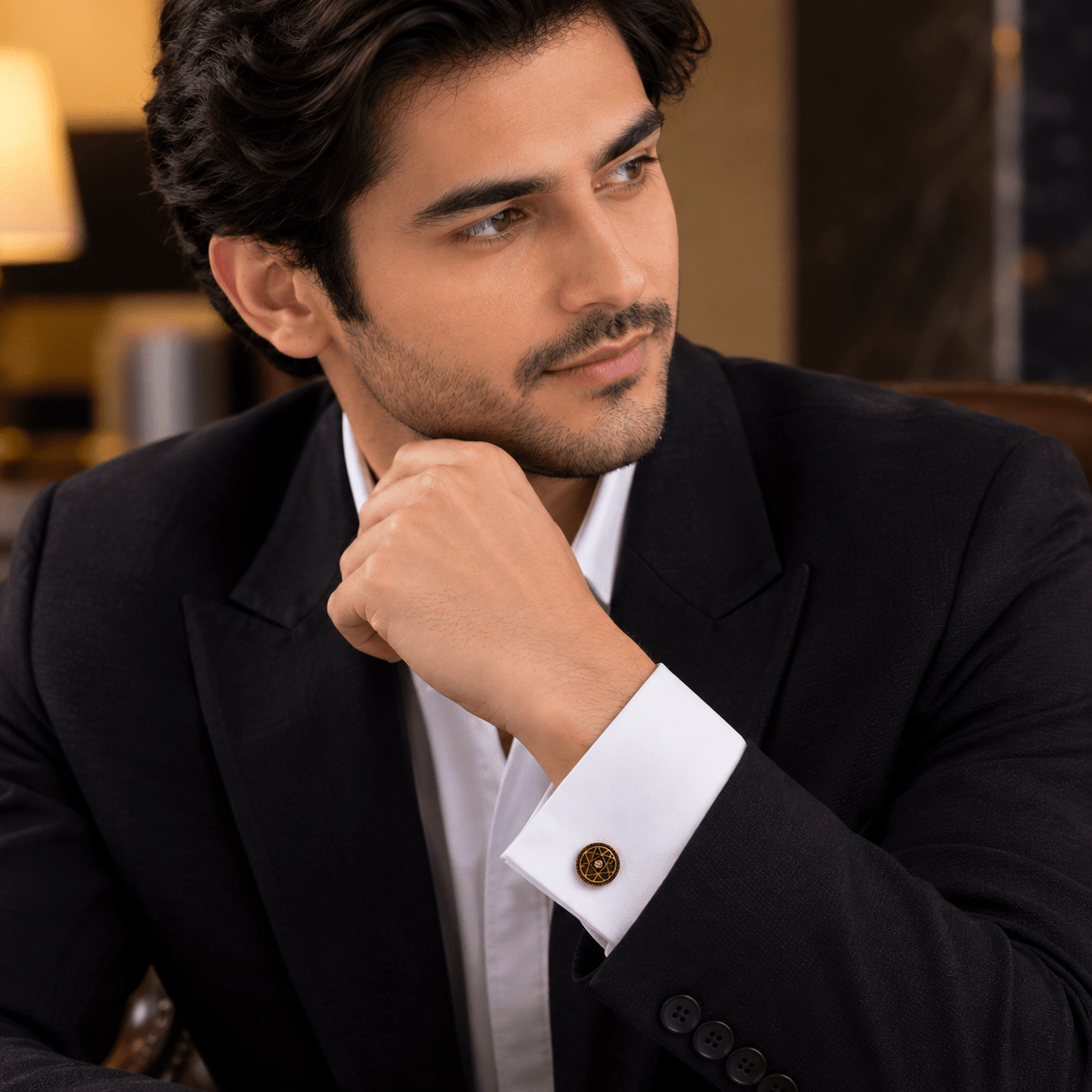A man in a black suit and white shirt resting his chin on his hand, showcasing a single gold button with a decorative circular emblem on the white cuff.