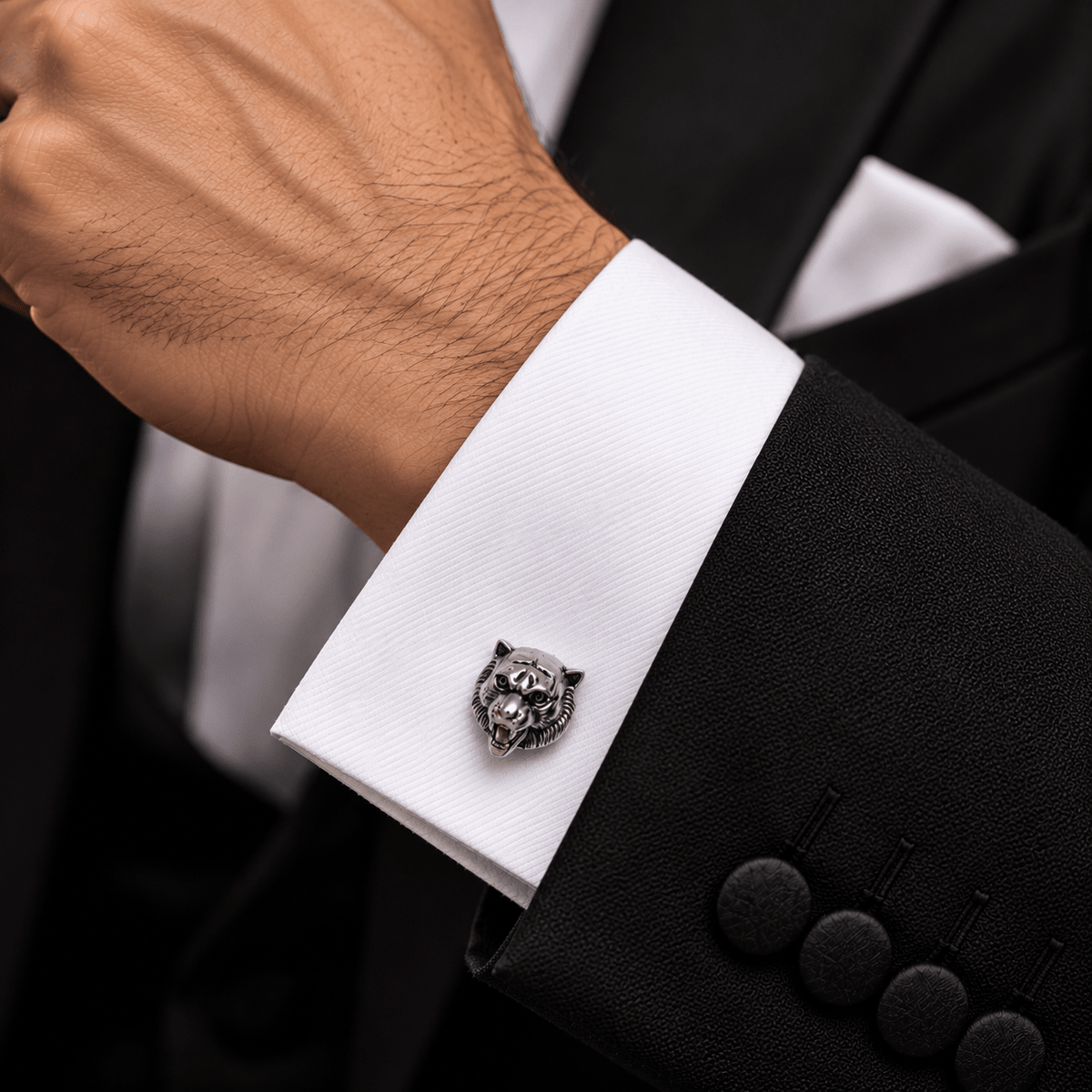 A close-up of a man's wrist wearing a white dress shirt cuff with a detailed silver tiger head cufflink, by Miraura Jewels paired with a black suit jacket.