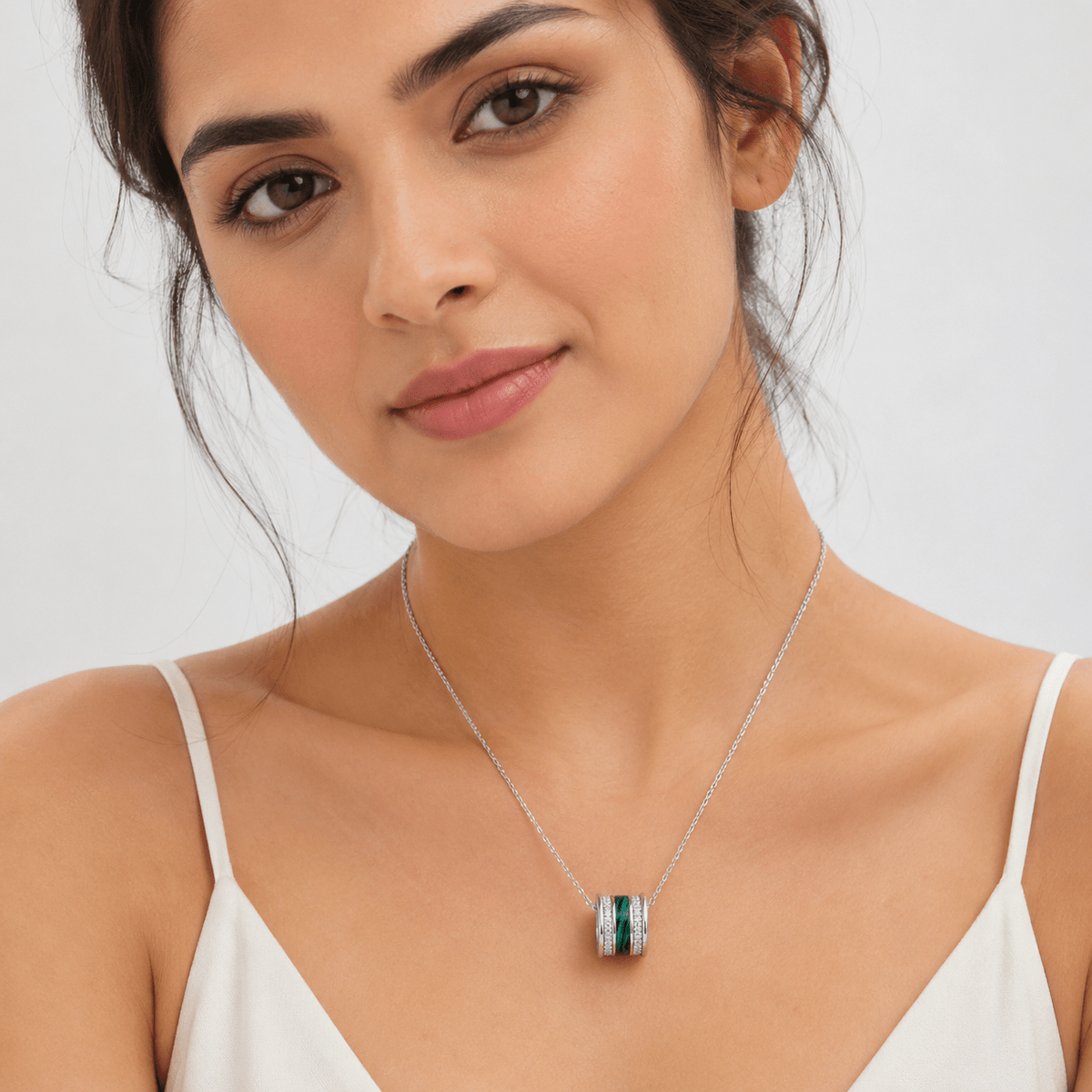 A close-up of a woman wearing a Silver Malachite Pave Bead Pendant, by Miraura Jewels. Silver-toned necklace with a cylindrical pendant featuring a vertical green malachite inlay flanked by small clear stones.
