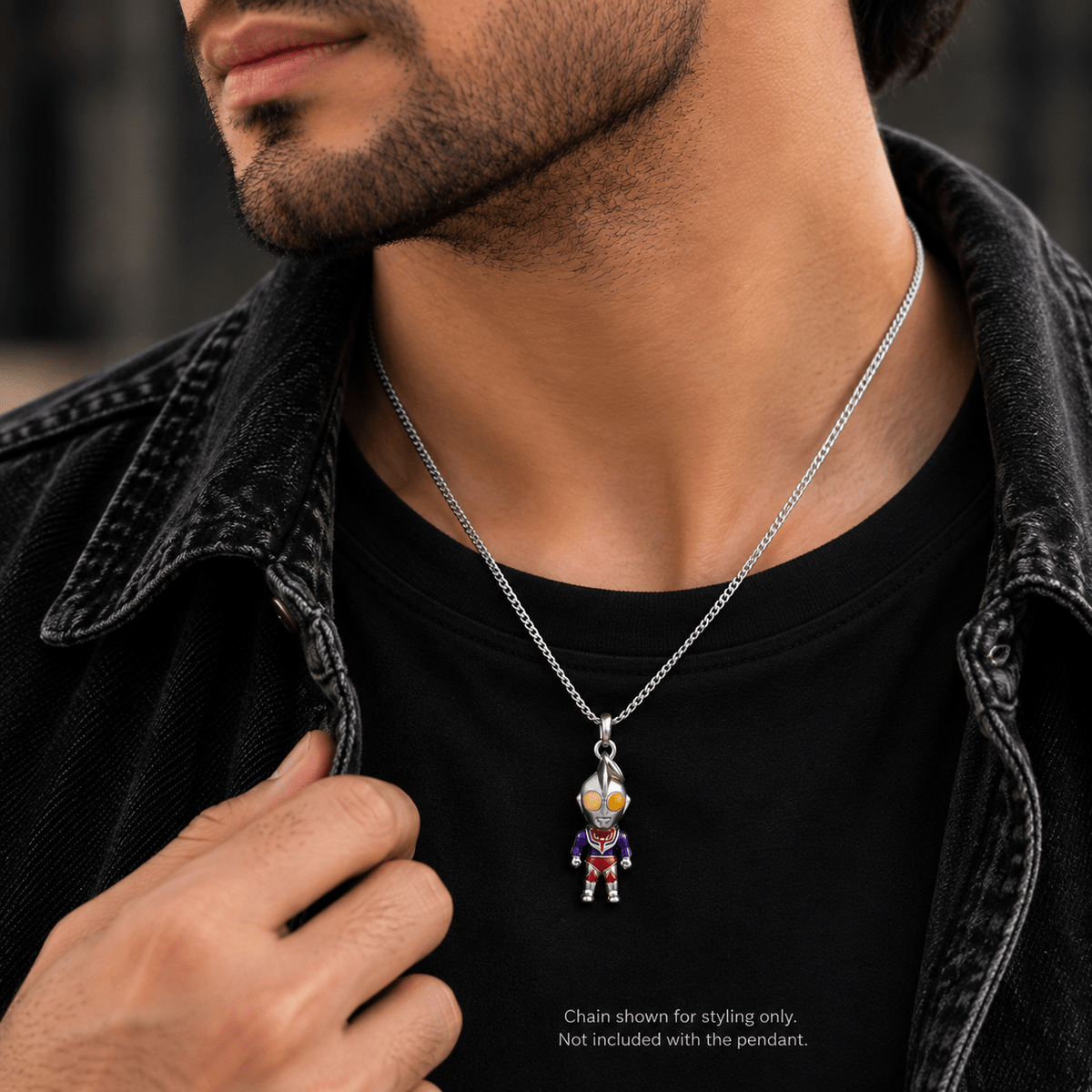 A close-up of a man wearing a silver chain necklace with a small Ultraman-style character pendant, styled over a black t-shirt and dark denim jacket.