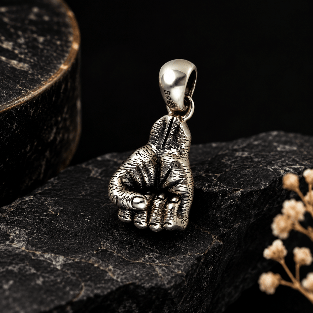 A sterling silver pendant shaped like a clenched fist, displayed on a dark textured stone surface with a dark background and small dried flowers.