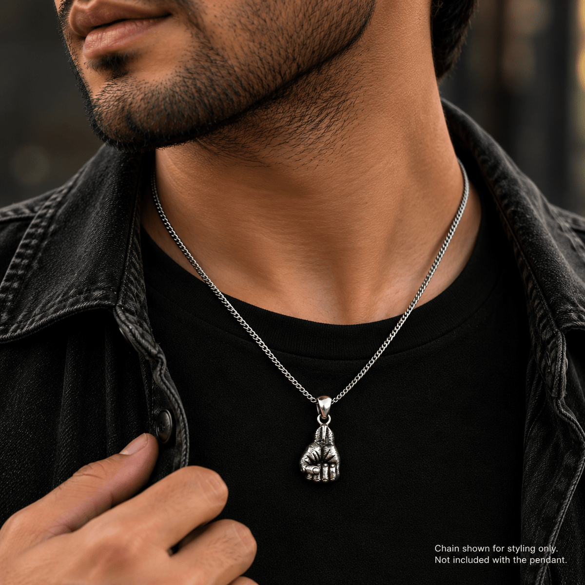 A close-up of a man wearing a silver chain necklace with a detailed fist pendant, styled over a black t-shirt and a dark denim jacket.
