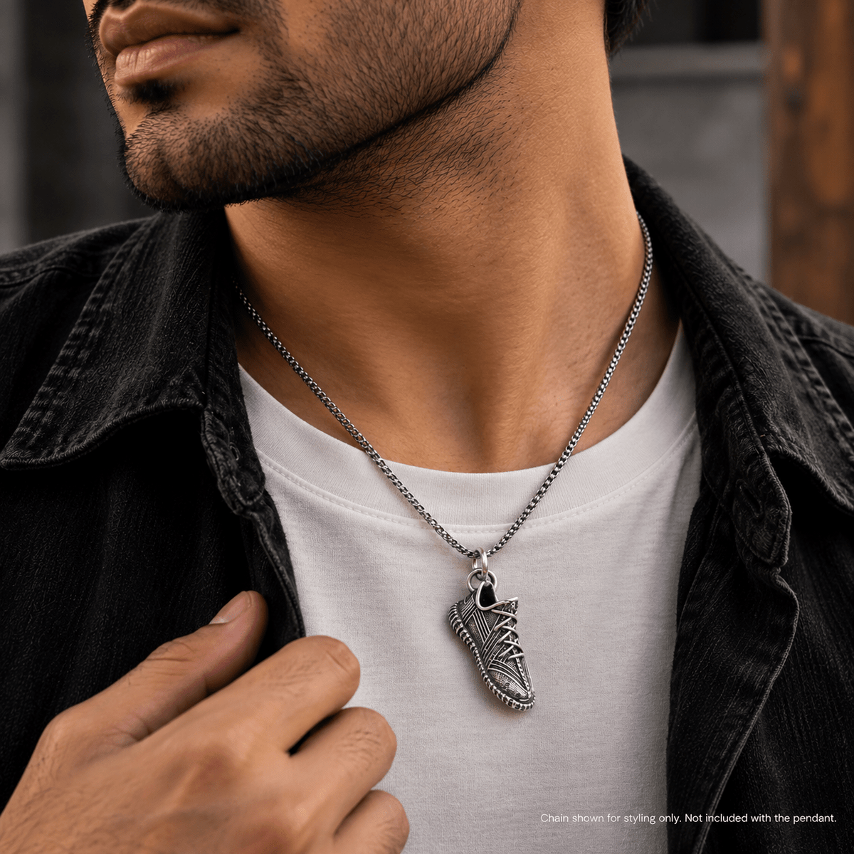 A close-up of a man wearing a silver chain necklace with a detailed sneaker-shaped pendant, styled over a white t-shirt and a dark denim jacket.