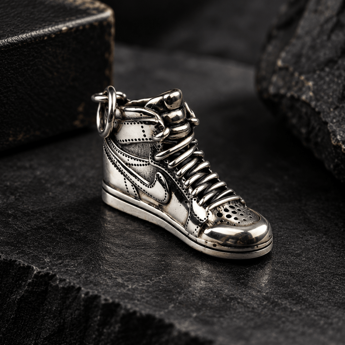 A metallic silver high-top sneaker  pendant with a key ring, photographed on a dark textured surface with a blurred dark box in the background.