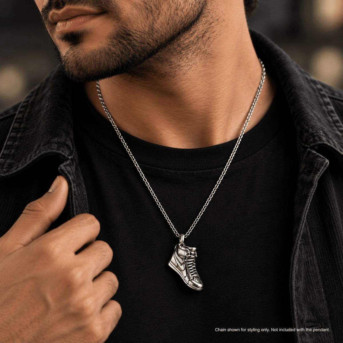 A silver-toned metal pendant shaped like a sneaker, worn on a chain by a man in a black t-shirt and dark denim jacket.