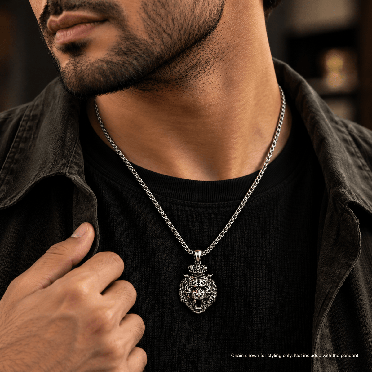 A close-up of a man wearing a silver chain necklace with an intricately detailed lion head pendant, styled over a black t-shirt and dark jacket.