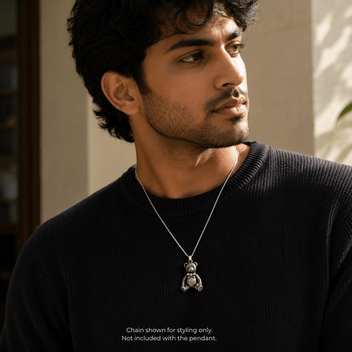 A man wearing a silver chain with a 925 silver teddy bear pendant, styled over a black knit sweater in warm natural light.