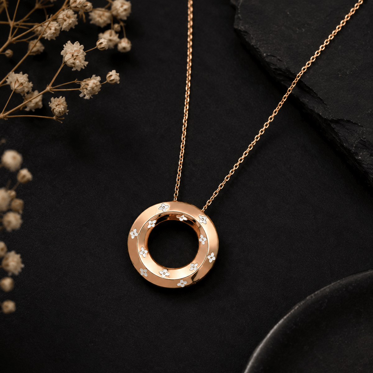 A Miraura's Rose Gold Segment Pendant displayed on a dark textured surface with dried baby's breath flowers.