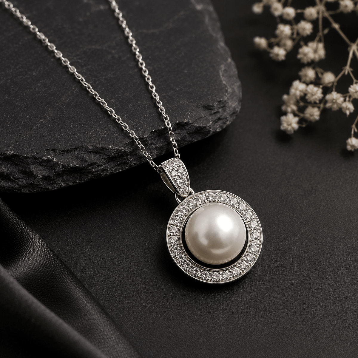 A silver pendant necklace featuring a large white pearl surrounded by a halo of small clear stones, displayed on a dark textured surface with a black stone and white dried flowers.