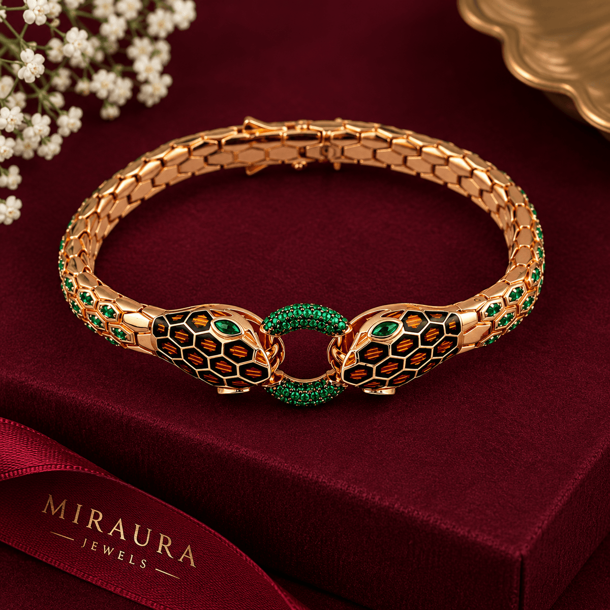 A Miraura Jewels Double  Headed Serpent Bangle, gold-plated snake-head bracelet with green and orange enamel details, displayed on a dark red velvet jewelry box with a ribbon.