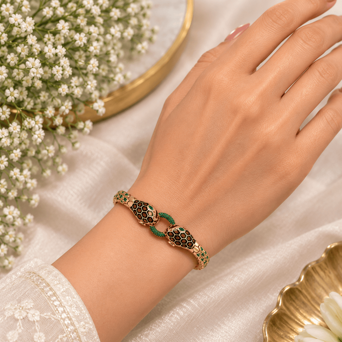 A Double Headed Serpent Bangle - by Miraura Jewels. Rose Gold-plated Snake-link bracelet with green and black enamel details, worn on a woman's wrist. The background features a soft white fabric, delicate white flowers, and gold-toned decorative plates.