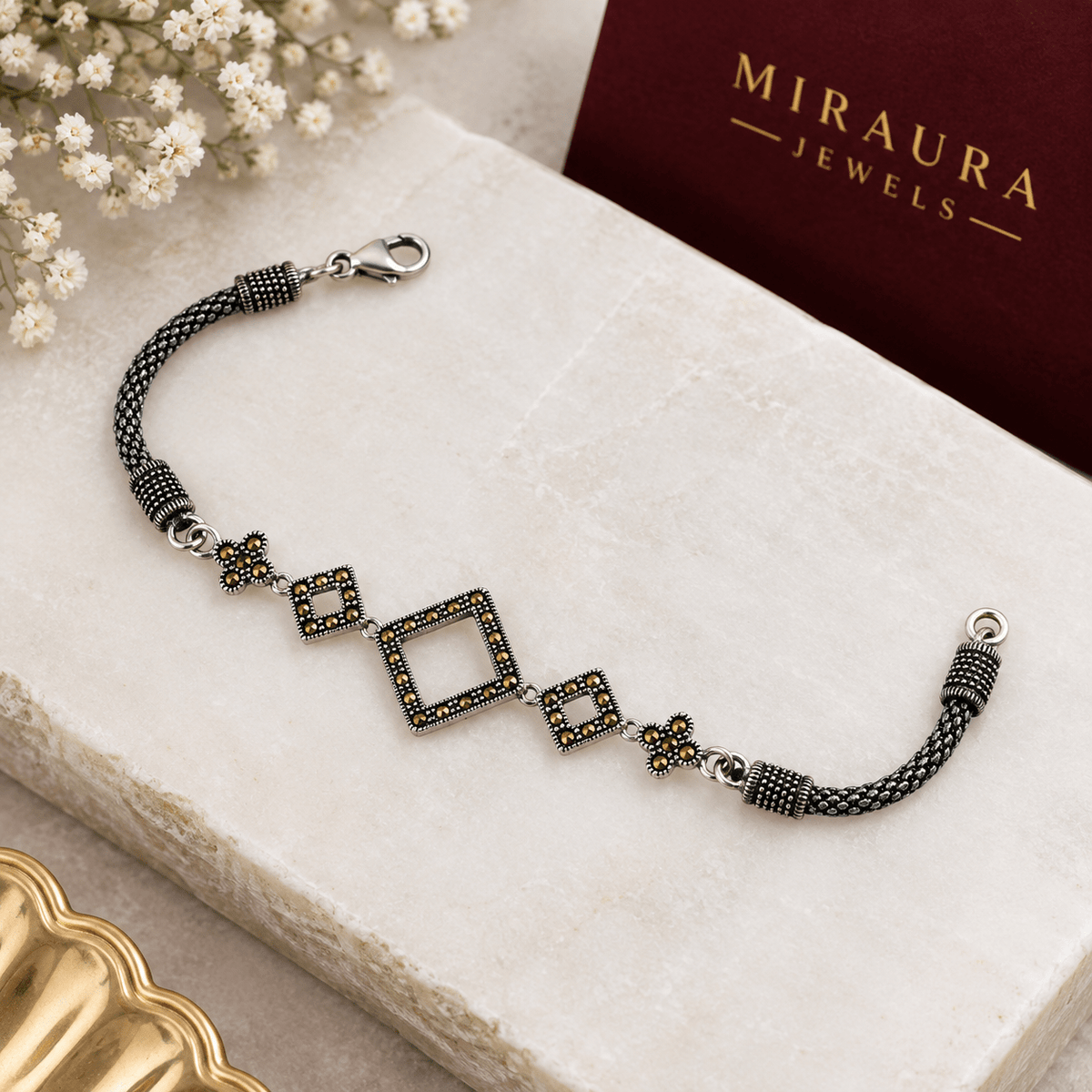 A Silver Tone Marcasite Kite Station Bracelet - by Miraura Jewels. Oxidised Silver Beaded bracelet with geometric square and floral motifs, featuring dark gray and black beads with small yellow accents, displayed on a textured cream stone slab next to a dark red Miraura Jewels box.