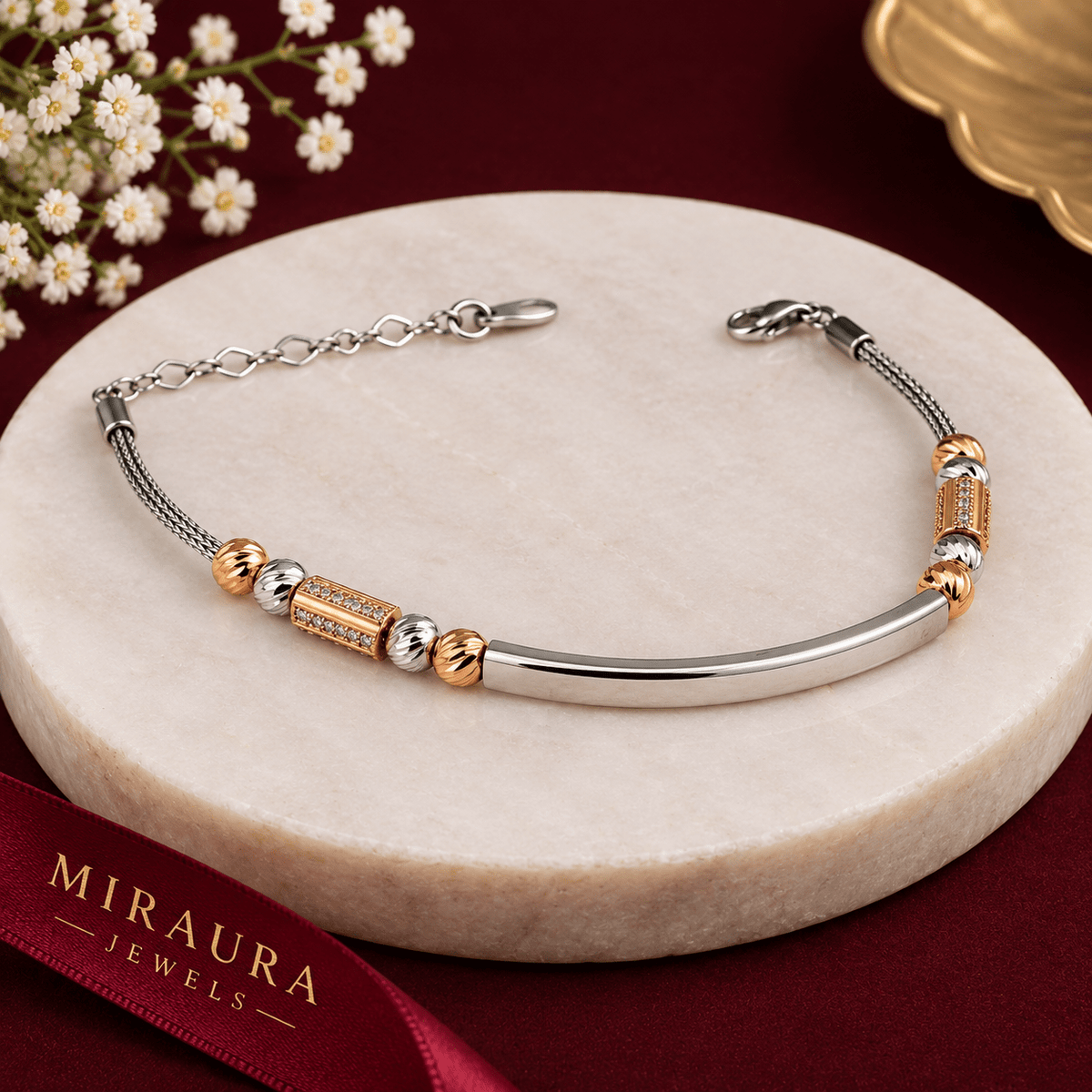 A silver and rose gold Tube and Bead Bracelet - by Miraura Jewels with a curved bar, textured cylindrical beads, and small clear stones, displayed on a round beige stone pedestal. A red ribbon with 'MIRAURA JEWELS' branding is visible in the foreground, with small white flowers in the top left corner.