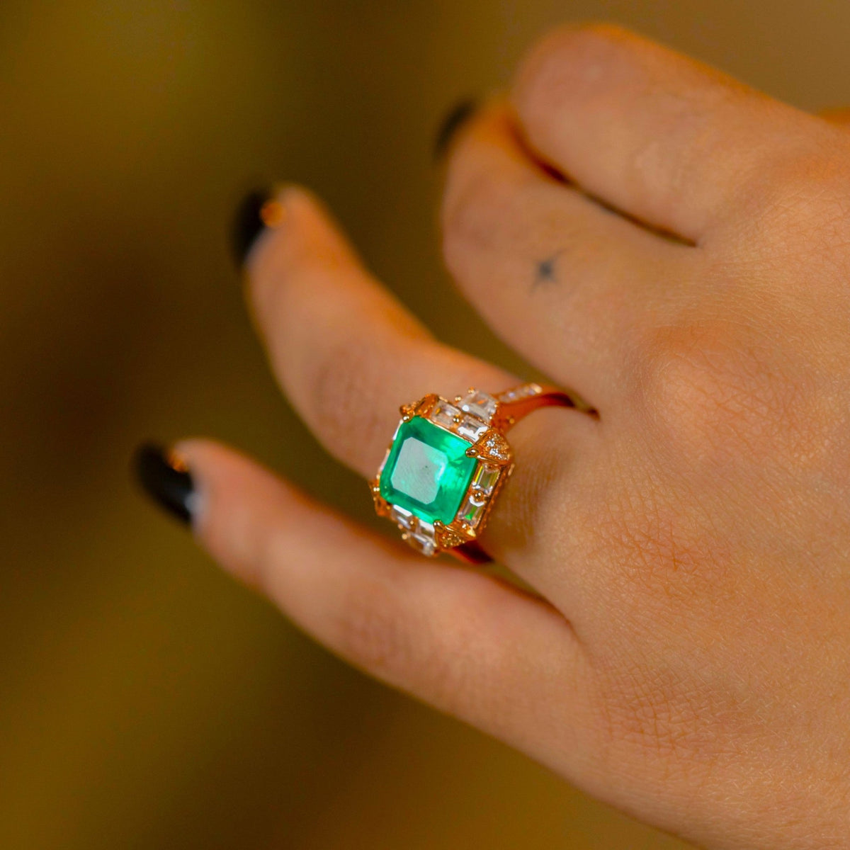 A close-up of a hand wearing a ring with a large square-cut green emerald gemstone in a gold setting, surrounded by small clear stones.