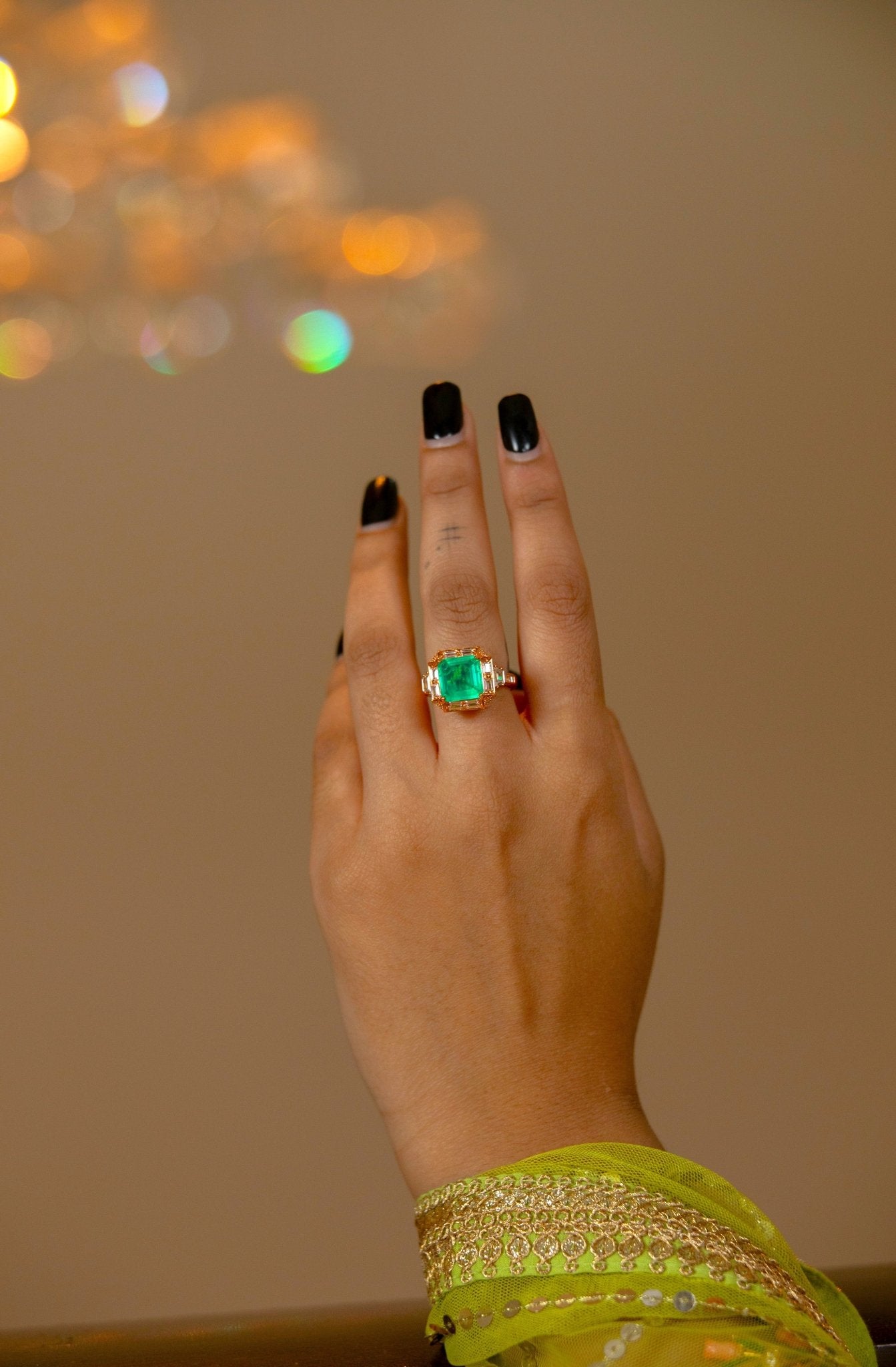  Hand wearing Miraura's Emerald Green Statement Ring with a blurred chandelier in the superbly lit background.