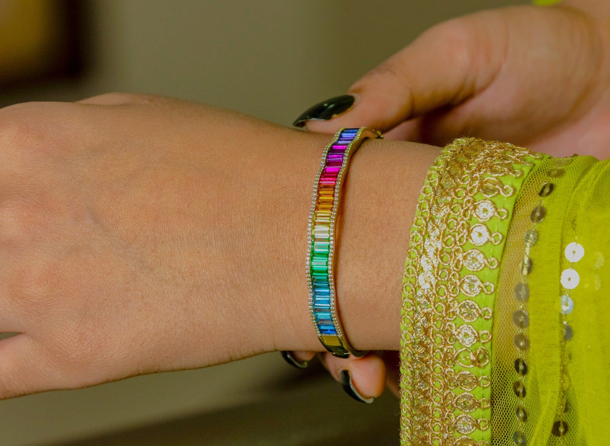 A close-up of a person's wrist wearing a slim metallic bracelet with a vibrant rainbow of rectangular gemstones. The person is also wearing a lime green embroidered sleeve.